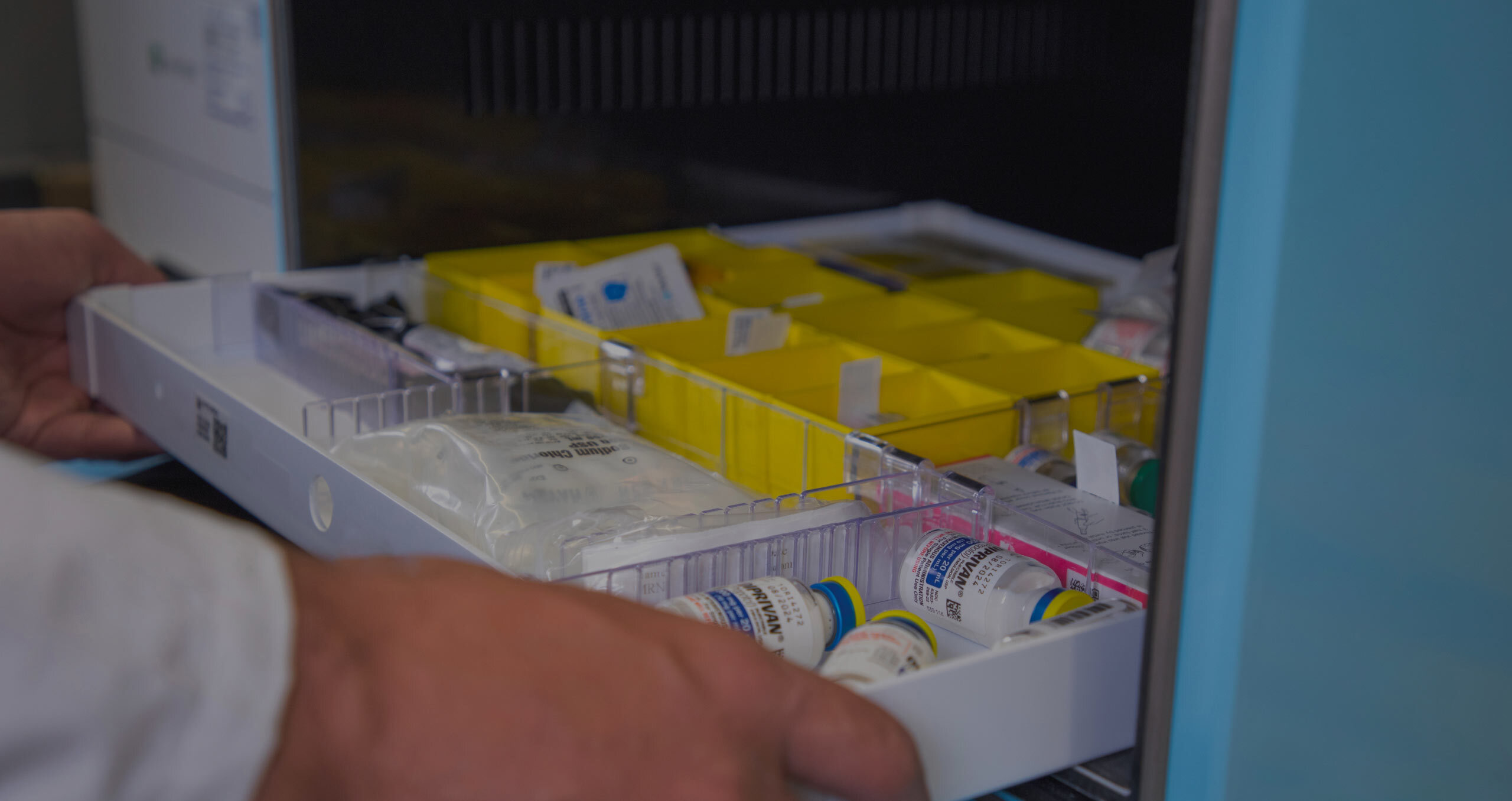 A hand reaches for a tray of vials in a storage unit, with yellow compartments and various medical supplies visible.